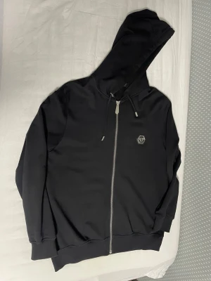 Svart hoodie från Philipp Plein - Säljer en svart hoodie från Philipp Plein med dragkedja och huva. Framsidan har en liten logga på bröstet och baksidan har en stor, detaljerad döskalle i vitt och grått samt Philipp Plein-tryck. Hoodien har ribbade muddar och snörning i huvan.
