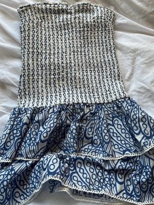 Blåvit axelbandslös kortklänning - Supersöt axelbandslös kortklänning i blått och vitt med smockad överdel och volangig kjol. Klänningen har ett unikt mönster med blå cirklar och linjer samt dekorativ spetskant längst ner. Perfekt för sommaren.