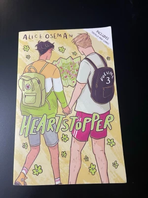 Heartstopper 3 - Heartstopper Volym 3 är en grafisk roman med färgglad illustrerad framsida som visar två killar som håller varandra i handen. Boken är på engelska och har ett mjukt omslag med tecknade detaljer och blommor. Perfekt för dig som gillar romantik och vänskap.