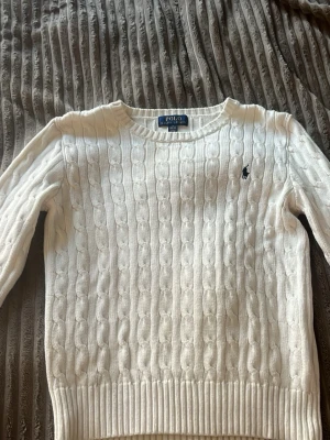 Vit kabelstickad tröja Polo Ralph Lauren - Kabelstickad vit tröja från Polo Ralph Lauren med rund halsringning och den klassiska broderade loggan på bröstet. Tröjan har ribbade muddar vid ärmslut och nederkant och är tillverkad i mjukt material. Perfekt för en clean och stilren look.