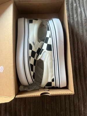 Vans Slip-On Checkerboard sneakers - Klassiska Vans Slip-On sneakers med svartvitt checkerboard-mönster. Skorna har låg profil, vit sula med svart rand och är tillverkade i canvas. Perfekta för en avslappnad och cool stil.