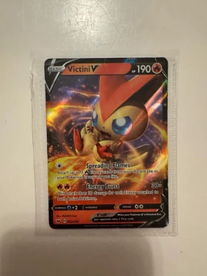 Victini V Pokémonkort - Victini V är ett samlarkort från Pokémon TCG med färgstark holografisk design. Kortet har 190 HP och attacker som Spreading Flames och Energy Burst. Det är i standardstorlek och har en mix av rött, gult och svart i motivet. Perfekt för dig som gillar att samla eller spela Pokémonkort.