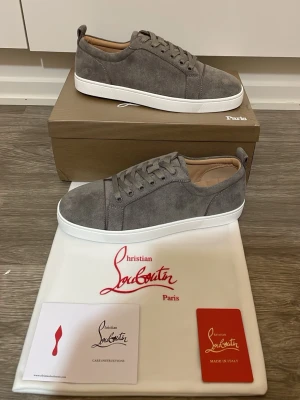 Christian Louboutin skor - Snygga grå sneakers från Christian Louboutin i mjuk mocka med vita platta sulor och klassisk snörning. Skorna har en stilren och minimalistisk design som passar perfekt till streetwear och vardagsstil.