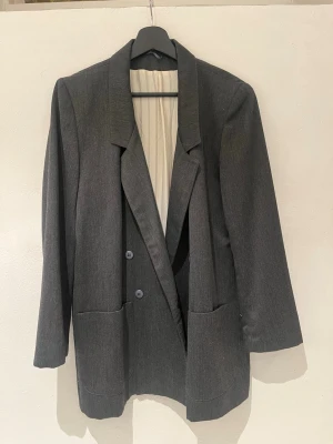 Grå oversized kavaj med fickor - Stilren grå kavaj med breda slag och dubbelknäppning. Modellen är oversized med två stora framfickor och långa ärmar. Materialet ser ut att vara en mjuk och lätt ullblandning. Perfekt för att skapa en cool och avslappnad look.