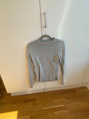 Ljusgrå stickad tröja med slits - En stilren ljusgrå stickad tröja med rund hals och ribbade muddar. Tröjan har långa ärmar och en liten slits i sidan nedtill för extra detalj. Perfekt att matcha med jeans eller kjol för en enkel och snygg look.