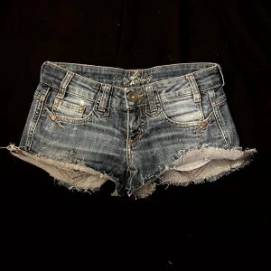 Lågmidjade jeansshorts  - Säljer ett par blå denim shorts med råa, fransiga kanter och klassiska fem fickor. Shortsens slitna look och låg midja ger en cool och avslappnad vibe. Perfekt för dig som gillar en edgy stil. Stängs med knapp och dragkedja. Stolek w26 och midjemått 77 cm