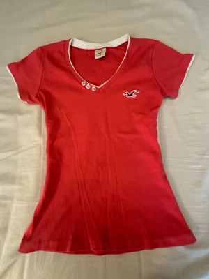 Röd v-ringad t-shirt från Hollister - Snygg röd t-shirt från Hollister med vit kant vid hals och ärmslut. T-shirten har v-ringning med tre dekorativa knappar och Hollisters logga broderad på bröstet. Modellen är figurnära och kortärmad, perfekt för en avslappnad stil.