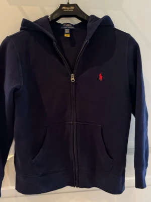 Mörkblå hoodie från Polo Ralph Lauren - Mörkblå hoodie med dragkedja från Polo Ralph Lauren. Klassisk design med huva, kängurufickor och det ikoniska röda Polo-logotrycket på bröstet. Tillverkad i mjuk bomullsblandning som känns skön mot huden. Perfekt för en avslappnad och stilren look.