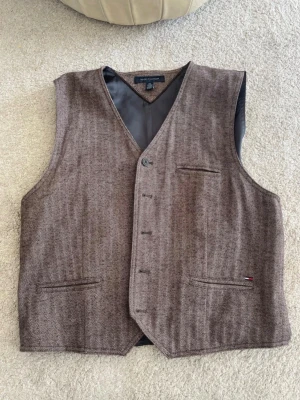 Brun ullväst från Tommy Hilfiger - Snygg brun väst från Tommy Hilfiger i klassisk v-ringad modell. Västen har diskret fiskbensmönster, tre fickor fram och knäppning med fyra knappar. Tillverkad i mjuk ullblandning och har ett elegant innerfoder. Perfekt för att lyfta din outfit.