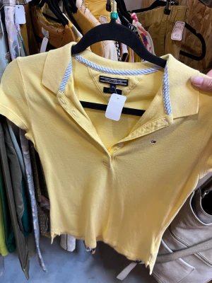 Gul pikétopp från Tommy Hilfiger - Snygg gul pikétopp från Tommy Hilfiger med klassisk krage och korta ärmar. Toppen har små knappar framtill och en diskret logga på bröstet. Tillverkad i mjuk bomull och har en vit-randig detalj på insidan av kragen.