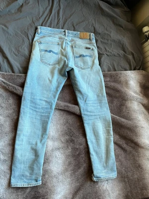Ljusblå jeans från Nudie Jeans - Säljer ett par ljusblå jeans från Nudie Jeans med klassisk femficksmodell och snygga kontrastsömmar. Jeansen har en slim fit passform och är tillverkade i mjuk bomullsdenim. Bakfickorna har Nudie Jeans ikoniska broderi och läderpatch i midjan.