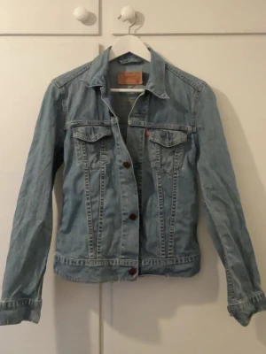 Levi's ljusblå jeansjacka S - Klassisk ljusblå jeansjacka från Levi's med raka sömmar, bröstfickor med lock och knappstängning framtill. Jackan har den ikoniska röda Levi's-taggen på bröstfickan och snygga kontrastsömmar. Perfekt att slänga över en hoodie eller t-shirt.