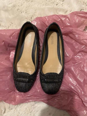 Svarta ballerinaskor från Michael Kors - Stilrena svarta ballerinaskor från Michael Kors med diskret logomönster och metallbricka framtill. Skorna har rundad tå och är tillverkade i ett tåligt konstläder med beige innersula för extra komfort. Perfekta för en trendig och klassisk look.