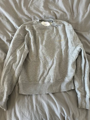 Grå sweatshirt från Axel Arigato - Säljer en stilren grå sweatshirt från Axel Arigato med gul broderad logga på bröstet. Tröjan har rund halsringning, ribbade muddar och är tillverkad i mjuk bomullsblandning. Perfekt för en avslappnad och trendig look.