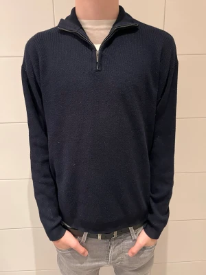 Mörkblå halvzip tröja - Mörkblå stickad tröja med halvzip och hög krage. Tröjan har långa ärmar och ribbade muddar vid ärmslut och nederkant. Perfekt för lager på lager och enkel att matcha med jeans eller chinos.
