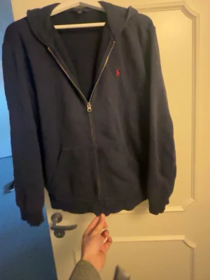 Marinblå hoodie från Polo Ralph Lauren - Snygg marinblå hoodie från Polo Ralph Lauren med dragkedja framtill och klassisk röd logga på bröstet. Hoodien har huva, fickor på magen och är tillverkad i mjukt material som passar perfekt till chill vardagsstil.