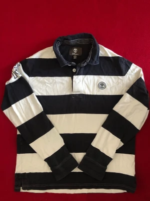 Randig rugbytröja från Timberland - Säljer en svartvit randig rugbytröja från Timberland Earthkeepers. Tröjan har långa ärmar, krage och knappar vid halsen. Tillverkad i bomull och har en broderad Timberland-logga på bröstet samt tryck på ena ärmen.