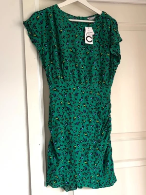 Grön leopardmönstrad klänning från C - Grön kortklänning från C med leopardmönster i blått, svart och gult. Klänningen har korta ärmar och en avslappnad passform. Perfekt för dig som vill sticka ut med färg och mönster.
