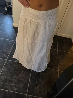 Vit långkjol med broderade blommor - Säljer en vit långkjol med bred smock i midjan och broderade blommor. Kjolen har volangdetaljer och är gjord i ett lätt bomullstyg som passar perfekt till varmare dagar. Superfin att styla med crop tops eller linnen.