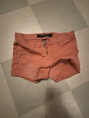 coral färgade shorts från billabong - Snygga coral shorts från billabong med rå kant nertill och klassisk femficksdesign. Shortsen har knapp och dragkedja framtill samt bälteshällor. Perfekta för varma dagar och har en tajt passform.