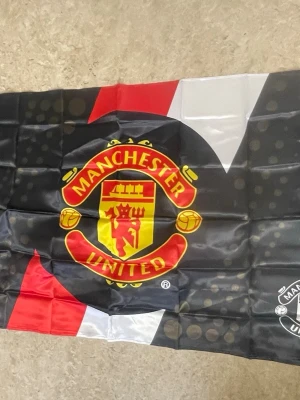 Manchester United supporterflagga - Stor supporterflagga med Manchester Uniteds klassiska klubbmärke i rött, gult och svart. Flaggan har en modern design med svarta, röda och vita fält och är tillverkad i ett tunt, glansigt syntetmaterial. Perfekt för att visa vilket lag du hejar på!