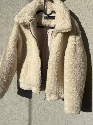 Krämvit teddyjacka från Zara - Mysig teddyjacka från Zara i krämvit färg med fluffig struktur. Jackan har hög krage, dragkedja framtill och är helfodrad. Passar till allt och är superenkel att styla.