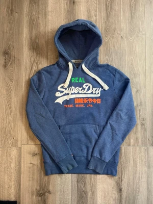 Blå Superdry hoodie med tryck - Blå hoodie från Superdry med stor vit logga och färgglatt tryck på bröstet🙌🏻 True to size alltså M😇 Hör av er vid funderingar samt snabbaffär🤗