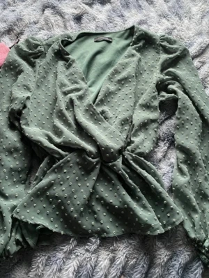 Grön omlottblus med prickig struktur - Stilren grön omlottblus med lång ärm och markerad midja. Blusen har en prickig struktur i tyget och lätt puffade axlar för en trendig look. Perfekt att matcha med jeans eller kjol för en snygg outfit.