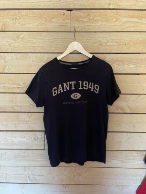 Marinblå GANT 1949 t-shirt - Snygg marinblå t-shirt från GANT med trycket 'GANT 1949' och 'New Haven Connecticut' på bröstet. Klassisk rund halsringning och korta ärmar. Perfekt till jeans eller shorts för en avslappnad look.