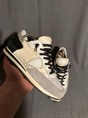 Philippe model  - Snygga sneakers från Philippe Model i beige, vitt och svart med svarta skosnören. Ovandelen är i en mix av mocka och textil, med detaljer i skinn. Klassisk låg modell med rund tå och kontrastfärgad häl. Perfekta för en trendig streetstyle-look.