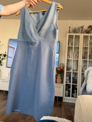 Ljusblå ärmlös klänning med omlott - Snygg ljusblå klänning med omlottdesign upptill och v-ringning. Överdelen har ett diskret randigt mönster och kjolen är slät. Klänningen är ärmlös och har dragkedja bak. Perfekt för dig som vill ha en stilren och elegant look.