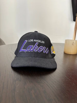 Svart Lakers keps Mitchell & Ness - Snygg svart keps med broderad Los Angeles Lakers-logga i lila och vitt framtill samt Lakers-märke på sidan. Kepsen har böjd skärm med lila undersida och justerbar baksida. Tillverkad i bomull och polyester, perfekt för dig som gillar basket och streetwear.