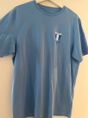 Ljusblå Malmö FF t-shirt - Snygg ljusblå t-shirt från Malmö FF med klubbens klassiska logga tryckt på bröstet. T-shirten har rund hals och korta ärmar, perfekt för dig som vill visa vilket lag du hejar på. Tillverkad i mjuk bomull för skön känsla. Helt ny med prislapp kvar 