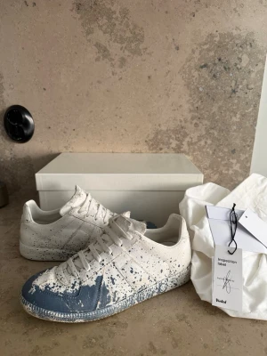 Maison Margiela blå-vita sneakers - Unika vita sneakers från Maison Margiela med blå färgstänk och splash-effekt över tå och sidor. Lågt skaft, klassisk siluett och vita skosnören. Tillverkade i läder med gummisula. Kommer med originalkartong och dustbag.  Skorna är stora i storlek passar upp till 401/2 Dom är äkta vill ni ha fler bilder är det bara säga till mvh MW