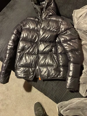 Exklusiv save the duck jacka  - Säljer en svart pufferjacka med glansig finish och huva. Jackan har två dragkedjeförsedda fickor framtill och breda, vadderade paneler. Märke syns på vänster ärm. Perfekt för kalla dagar och riktigt snygg streetstil.