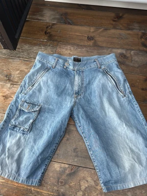 Blå jeansshorts från Energie - Säljer ett par blå jeansshorts från Energie med normal passform. Shortsen har flera fickor, inklusive en stor sidoficka med lock, och detaljerade sömmar. Klassisk denimlook med dragkedja och knappstängning. Perfekta för sommaren! Midja: 44 cm, längd: 63 cm, innerbenslängd: 33 cm  