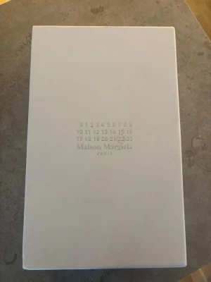 Maison Margiela vita sneakers - Stilrena vita sneakers från Maison Margiela med klassisk design. Skorna har ovandel i vitt läder och mocka, samt en kontrasterande brun gummisula. Perfekta för dig som gillar minimalistisk och tidlös stil.