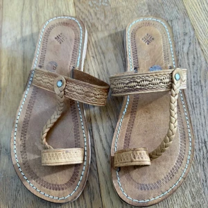 Handgjorda bruna lädersandaler - Unika sandaler i ljust brunt läder med flätad rem och dekorativa mönster på ovandelen. Tåremmen och den breda fotremmen ger en traditionell look. Sula i gummi för bra grepp. Perfekta för dig som gillar boho eller etno-stil.