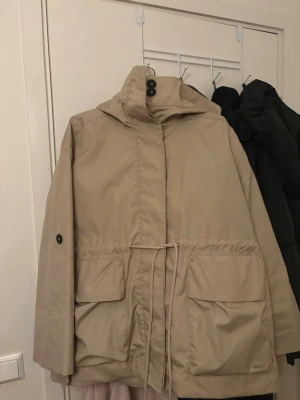 Beige parkasjacka från Everest - Stilren beige parkasjacka från Everest med huva, stora fickor framtill och dragsko i midjan. Jackan har en enkel design med knappar vid kragen och är tillverkad i ett slitstarkt material som passar perfekt för kyliga dagar. Har använts 2 gånger mycket bra skick ser ut som ny 
