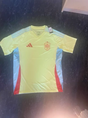 Spaniens fotbollströja Adidas gul - Gul fotbollströja från Adidas med Spaniens landslagsmärke på bröstet. Tröjan har ljusblå detaljer på axlar och sidor samt röda inslag. Tillverkad i ett lätt och ventilerande material, perfekt för träning eller match.