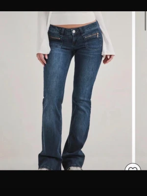 Mörkblå bootcut jeans med låg midja - Säljer ett par mörkblå bootcut jeans med låg midja och snygga detaljer som dragkedjor och dubbla fickor framtill. Jeansen har en klassisk tvätt och sitter tajt över höfterna innan de släpper vid benen. Perfekta för en trendig Y2K-look.