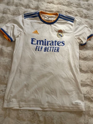 Real Madrid matchtröja Adidas vit - Vit Real Madrid matchtröja från Adidas med blå och gula detaljer. Tröjan har klubbmärke, Adidas-logga och 'Emirates Fly Better' tryckt på bröstet. Tillverkad i lätt funktionsmaterial med Aeroready-teknologi. Perfekt för fotbollsträning eller support.