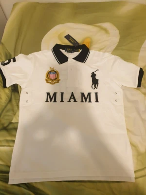 Vit Polo Ralph Lauren t-shirt MIAMI - Vit t-shirt från Polo Ralph Lauren med svart krage och detaljer. Stor broderad logga och texten MIAMI framtill samt emblem på bröstet. Klassisk pikédesign med korta ärmar och knappslå. Perfekt för en sportig och stilren look. Säljer på grund av att den inte passar mig. Har inte använts. Kan gärna diskutera priset.
