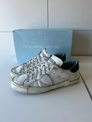 Philippe Model vita sneakers i läder - Snygga vita sneakers från Philippe Model i äkta läder med svarta detaljer vid hälen. Klassisk låg modell med vita snören och diskret logga på sidan. Perfekta för en stilren och trendig look. Innersulan är lagade som du ser på dem två sista bilderna.