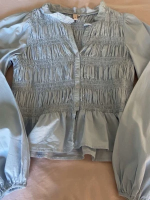 Ljusblå smockad blus med volang ifrån Nelly💓 - En ljusblå blus med smockad överdel och volang nertill. Blusen har långa puffärmar med resår vid ärmslut och knappar framtill. Den är i storlek 34 ifrån Nelly💓💓