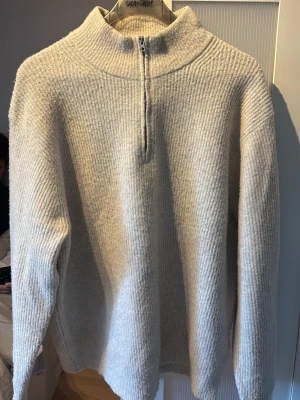 Beige stickad tröja från Uniqlo - Mysig beige stickad tröja från Uniqlo med halv dragkedja framtill och hög krage. Tröjan har en avslappnad passform och är tillverkad i ett mjukt, ribbat material.