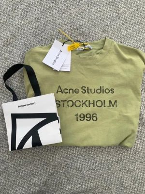 Grön Acne Studios t-shirt Stockholm - Snygg grön t-shirt från Acne Studios med trycket 'STOCKHOLM 1996' på bröstet. Klassisk rund hals och korta ärmar. Perfekt för dig som gillar stilrena plagg med en urban touch. Materialet är mjuk bomull och t-shirten har en normal passform.