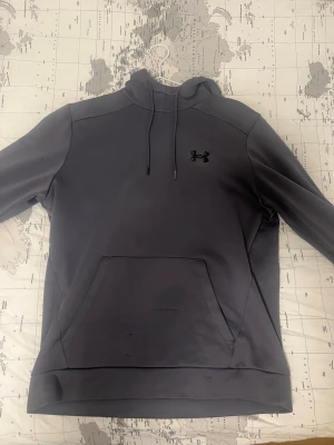 Mörkgrå hoodie från Under Armour - Snygg mörkgrå hoodie från Under Armour med svart logga på bröstet. Hoodien har dragsko i huvan och en stor känguruficka framtill. Tillverkad i mjukt och bekvämt material, perfekt för en avslappnad stil.