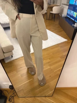 Beiga vida kostymbyxor - Snygga beiga kostymbyxor med hög midja och raka, vida ben. Byxorna har hällor för bälte och stängs med dragkedja och hake. Perfekta för en stilren och trendig look. Lite små fläckar som ni kan se på sista bilden, men inget som syns tydligt på. Finns matchande kavaj att köpa till separat om man önskar de.