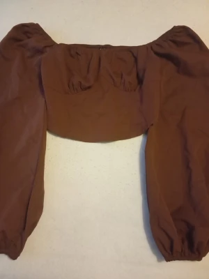 Brun offshoulder topp med puffärmar - Trendig brun offshoulder topp med långa puffärmar och smockad rygg. Toppen har en croppad passform och är perfekt för dig som vill sticka ut med en cool och avslappnad stil. Tillverkad i mjukt bomullstyg.